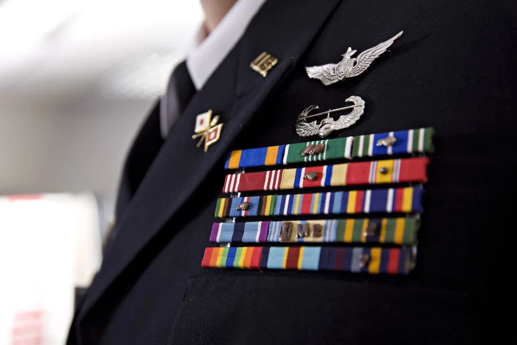 Close-up of a person in a military uniform displaying various service ribbons, medals, and pins on their chest, symbolizing achievements and adherence to strict compliance frameworks. The background is blurred.