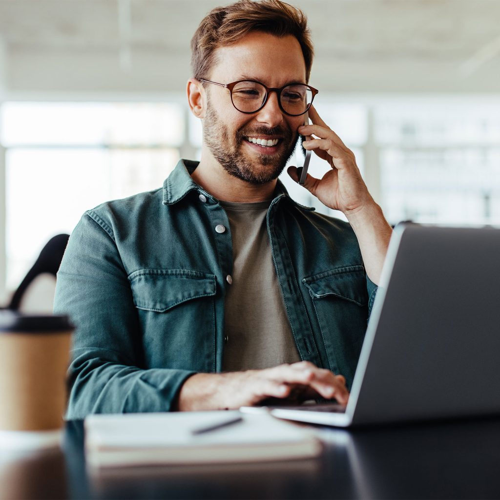 A smiling man with glasses talks on a smartphone while typing on a laptop at a desk, with a coffee cup and notebook nearby in a bright, modern office.