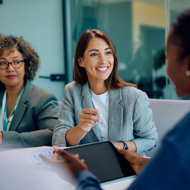 Three women sit at a table in a modern office, engaged in conversation. One woman in a gray blazer smiles and gestures as she shares her approach, while another listens attentively. The third woman holds a tablet.