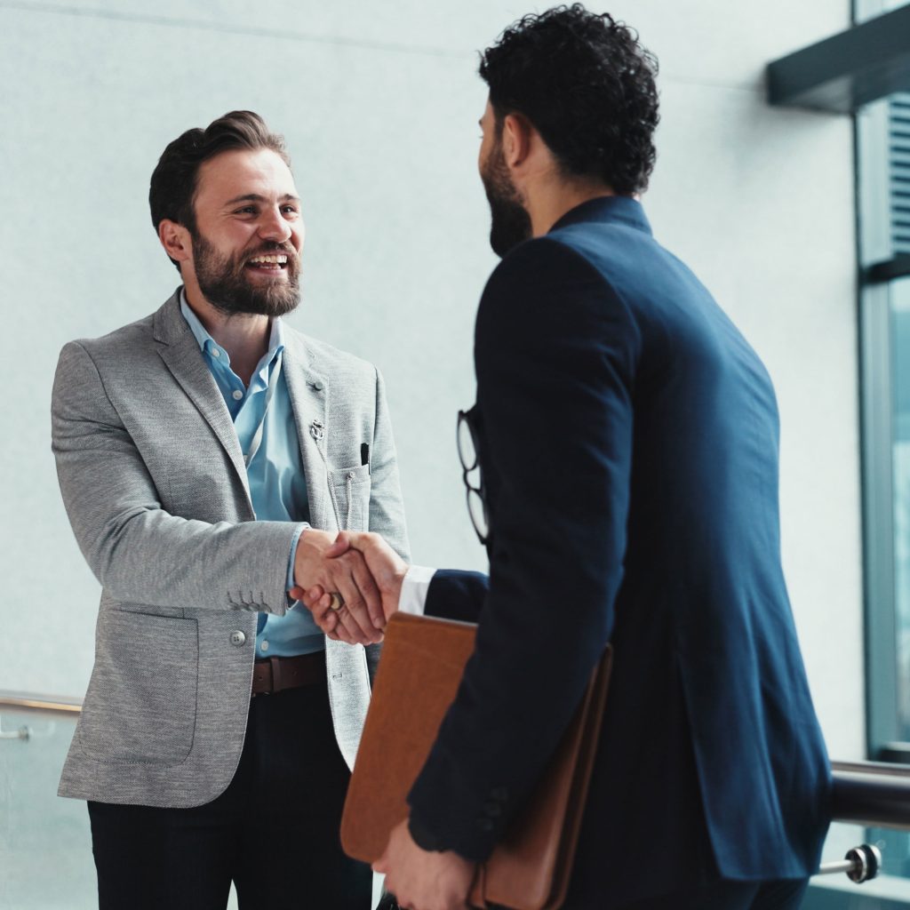 Two men in business attire shake hands and smile at each other in a modern office setting, suggesting a professional greeting or agreement—possibly a successful deal involving Ingram Micro. One holds a brown folder.