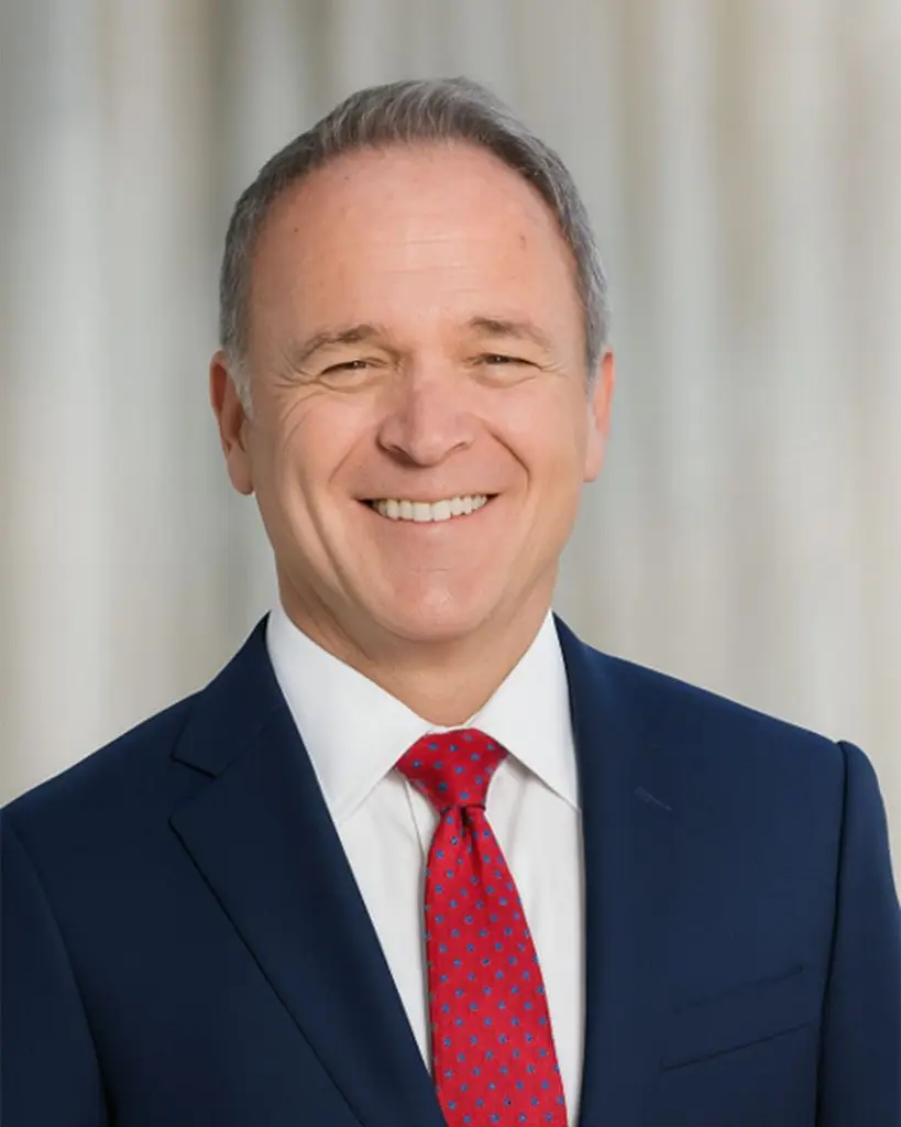 A smiling man in a blue suit, white shirt, and red patterned tie poses in front of a blurred light-colored background.