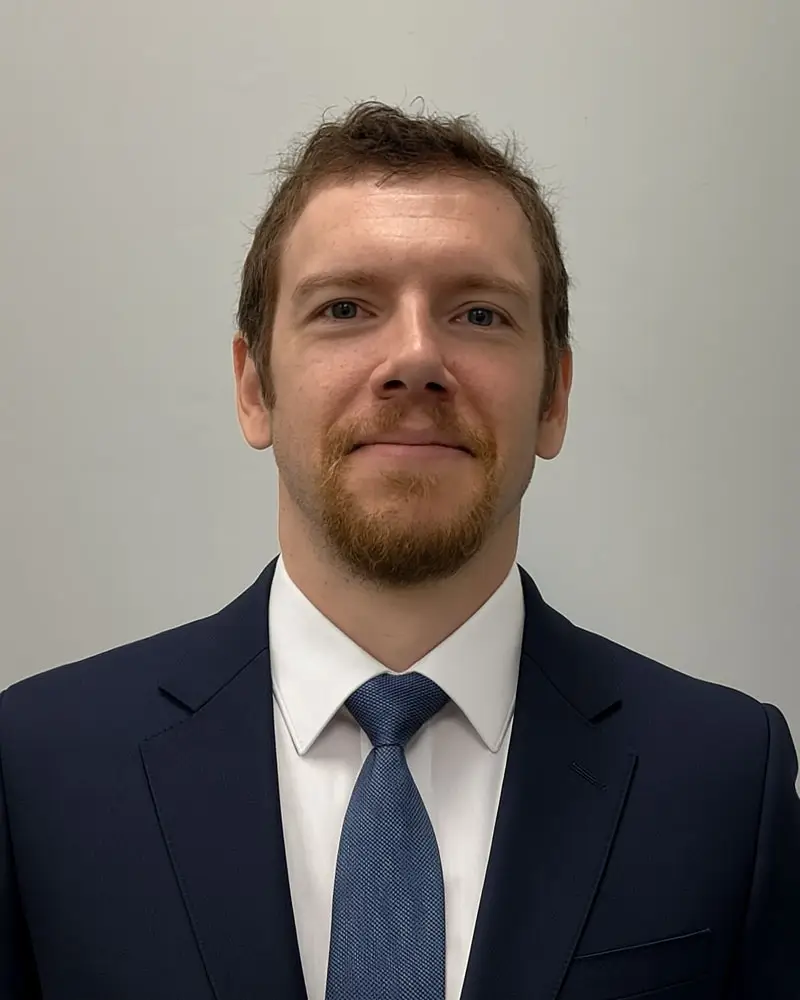 A man with short brown hair and a beard, wearing a dark suit, white shirt, and blue tie, stands against a plain light-colored background, facing the camera and smiling slightly.