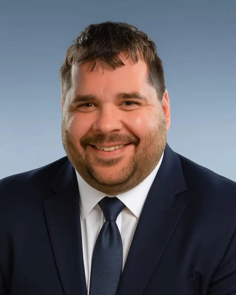 A man with short dark hair and a beard smiles at the camera, wearing a navy suit, white shirt, and navy tie, against a plain blue-gray background.