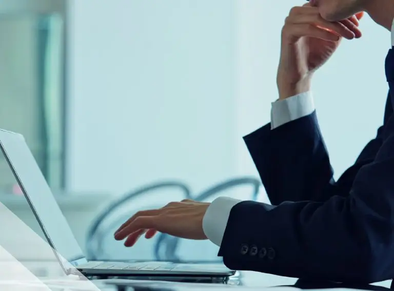 A person in a suit sits at a desk, resting their chin on one hand and typing on a FedRAMP Rev. 5-compliant laptop with the other. The background is blurred, showing a modern office setting.