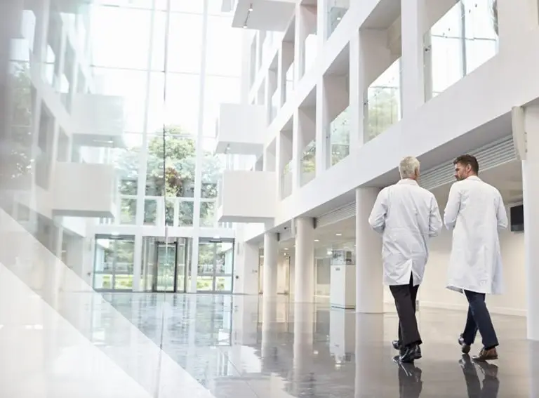 Two doctors in white lab coats walk and talk in a modern, bright hospital lobby with large glass windows, white walls, and shiny floors.