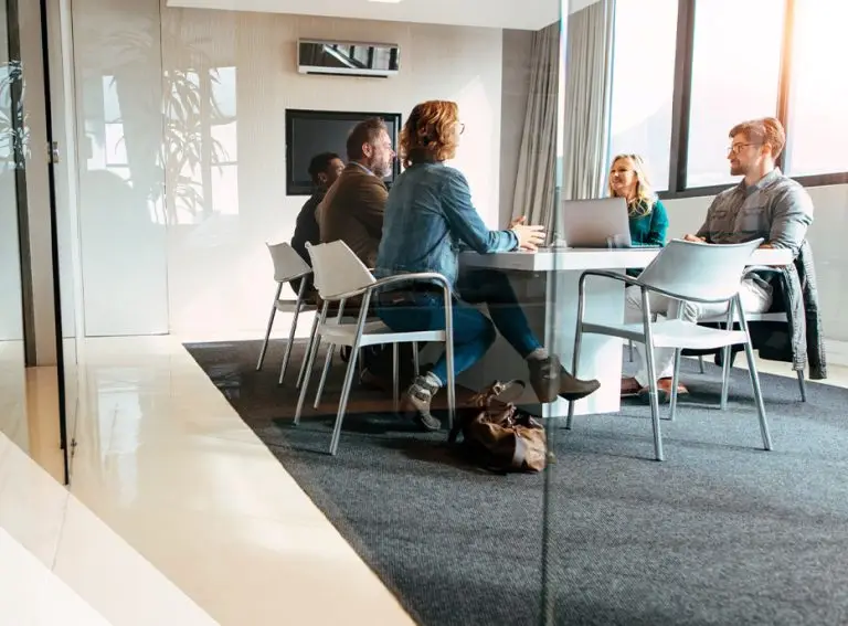 Five people sit around a conference table in a modern office, engaged in conversation about HITRUST compliance. Some have laptops open, and daylight streams through large windows. The professional atmosphere highlights their shared responsibility.