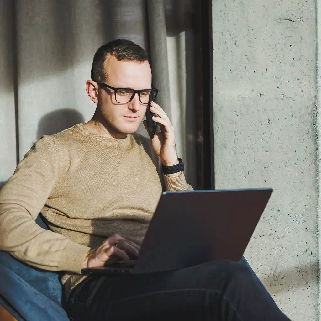 A man wearing glasses and a beige sweater sits by a window, talking on a smartphone while working on his laptop. Sunlight streams in, highlighting his focused expression as he discusses DoD compliance requirements.