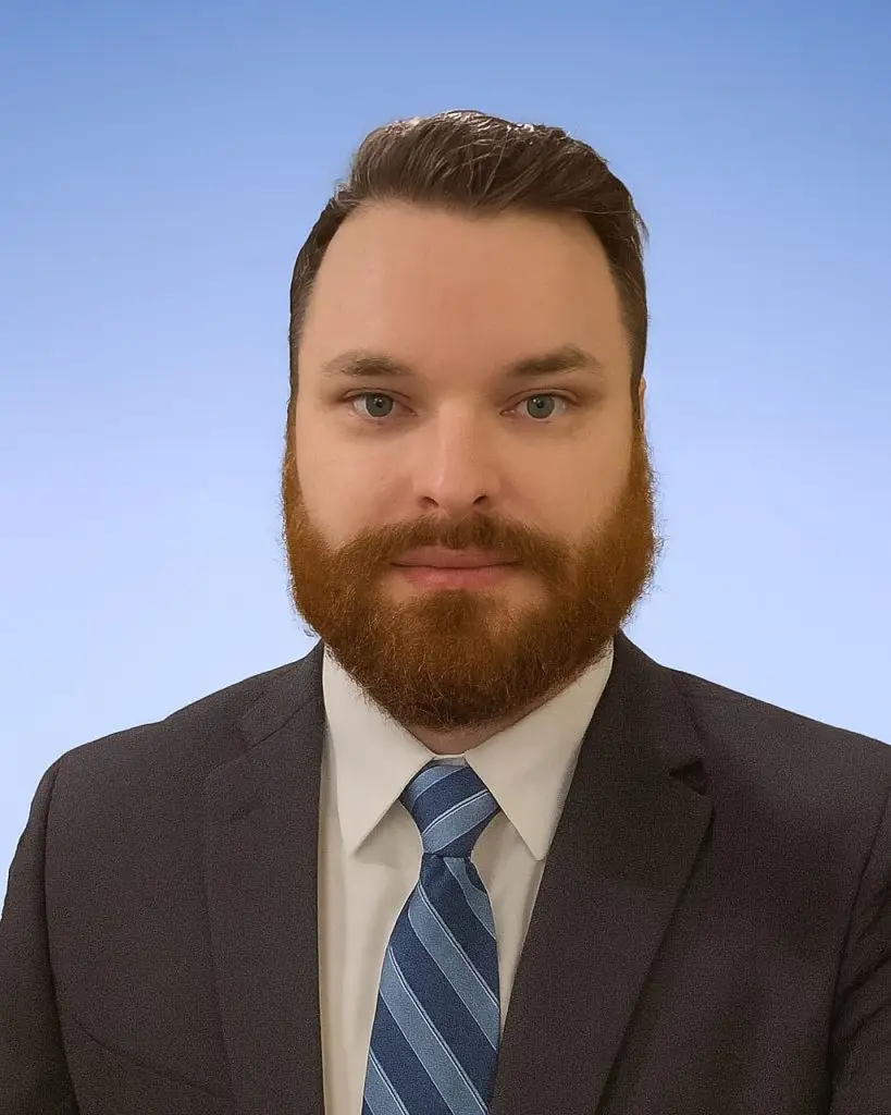 A man with a brown beard and short hair, wearing a dark suit, white shirt, and blue striped tie, poses against a light blue background.