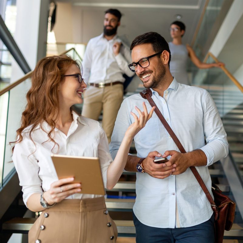 Two people wearing glasses smile and chat while walking down stairs in an office building, reflecting Our Approach to collaboration. The woman holds a tablet, the man a phone, with two others visible in the background also descending the stairs. Two people wearing glasses smile and chat while walking down stairs in an office building, reflecting Our Approach to collaboration. The woman holds a tablet, the man a phone, with two others visible in the background also descending the stairs.