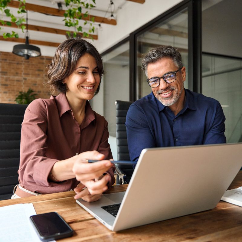 Two colleagues sit at a wooden desk, smiling and looking at a laptop screen together as they discuss a Microsoft Azure cloud computing project. Office plants and glass walls are visible in the background. Two colleagues sit at a wooden desk, smiling and looking at a laptop screen together as they discuss a Microsoft Azure cloud computing project. Office plants and glass walls are visible in the background.