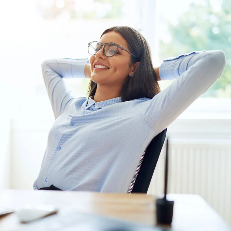 A woman wearing glasses and a light blue shirt leans back in her chair, smiling and relaxing with her hands behind her head at a desk in a bright office. A woman wearing glasses and a light blue shirt leans back in her chair, smiling and relaxing with her hands behind her head at a desk in a bright office.